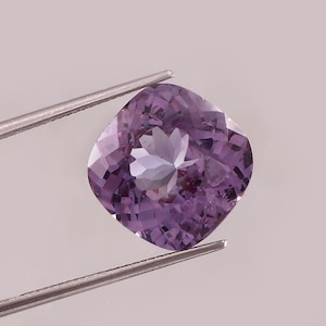 May include: A cushion-cut purple gemstone with a faceted surface, held by silver-toned metal tweezers. The stone displays deep violet hues and internal light reflections across its polished, symmetrical, and rounded square-shaped facets.