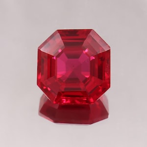 May include: A faceted, octagonal-cut ruby gemstone. The gem is a deep, vibrant red colour, with multiple facets that reflect light. The gemstone is sitting on a reflective surface.
