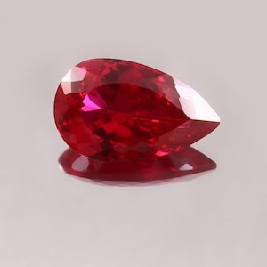 May include: A faceted, pear-shaped ruby gemstone. The gem is a deep, saturated red color with a high degree of clarity, reflecting light across its many facets. The stone is displayed on a reflective surface.