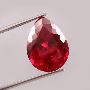 Peut inclure: Une pierre précieuse rouge foncé en forme de poire facettée est tenue par une pince argentée. La surface de la gemme reflète la lumière, mettant en valeur sa taille et sa couleur. La gemme est d'un rouge vif et saturé.