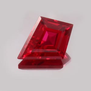 May include: A faceted, red gemstone with a kite-like shape. The gem has a square center and angled sides, reflecting light. The stone is displayed on a reflective surface, showcasing its brilliance. The gem is a deep, rich red color.
