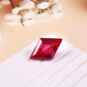 May include: A faceted, deep red gemstone with a geometric, kite-like shape. The gem is set against a white surface with a soft, blurred background of white and cream tones. The gem's facets reflect light, creating a brilliant display.