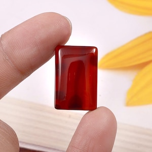 Puede incluir: Una gema rectangular y pulida con un tono rojo intenso, sostenida entre dos dedos. La piedra tiene una superficie brillante y parece translúcida, con vetas más oscuras visibles. El fondo incluye una flor amarilla borrosa.