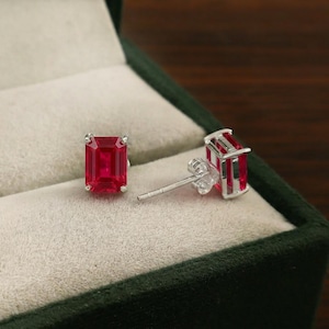 Può includere: Un paio di orecchini a bottone con gemme rosso rubino. Ogni orecchino presenta una pietra rossa vibrante a taglio rettangolare incastonata in una montatura in metallo color argento. Gli orecchini sono esposti in un portagioie verde scuro.