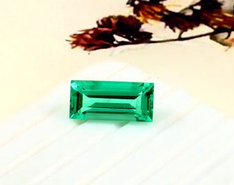 May include: A rectangular, emerald-cut gemstone with a vibrant green hue. The gem is set against a white surface, with a blurred background featuring floral elements. The gemstone's facets reflect light, showcasing its clarity and brilliance.