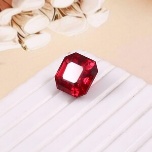 May include: A faceted, rectangular-cut, deep red gemstone with a white center. The gem is set against a white, ridged surface, with a soft, patterned background.