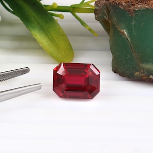 Peut inclure: Une pierre précieuse rouge foncé taillée en rectangle. La gemme est présentée sur une surface blanche, avec une paire de pinces argentées à gauche. Du feuillage vert et jaune et un morceau de roche verte et brune sont en arrière-plan.