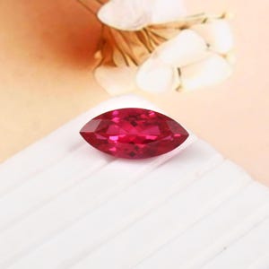 May include: A marquise-cut, faceted, ruby-red gemstone. The gem is set against a white, textured surface, with a soft, peach-coloured background. The gemstone's shape is elongated with pointed ends, showcasing its brilliant facets.