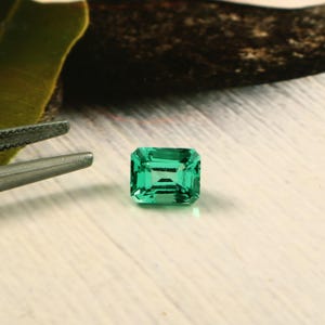Pode incluir: Uma gema vibrante, lapidação esmeralda, com um tom verde rico. A gema é retangular com uma superfície facetada, refletindo a luz. Uma pinça prateada é parcialmente visível no canto superior esquerdo. A gema está sobre uma superfície clara.
