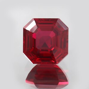 Puede incluir: Un rubí tallado en forma cuadrada. La gema es de color rojo intenso y saturado. La luz se refleja en las facetas, creando brillo. La gema se muestra sobre un fondo blanco, con su reflejo visible.