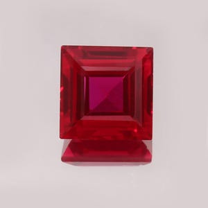 May include: A square-cut, faceted gemstone in a deep red colour. The gem features a central square within a larger square, creating a layered effect. The polished surface reflects light, enhancing its brilliance. The gemstone is likely for jewellery.