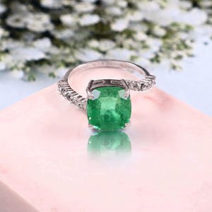 May include: A silver-toned ring featuring a large, cushion-cut green gemstone at the center, held by four prongs. The band is accented with a series of small, clear, round-cut stones along both sides, creating a bright and reflective appearance.