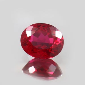 May include: A faceted, oval-cut ruby gemstone. The gem is a deep, vibrant red color with multiple facets that reflect light. The ruby is set against a white background, with its reflection visible below.