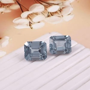 Puede incluir: Dos gemas azul claro de talla cuadrada con un diseño de talla escalonada. Las gemas se exhiben sobre una superficie blanca con un fondo suave y borroso con acentos florales blancos y un degradado de color melocotón.