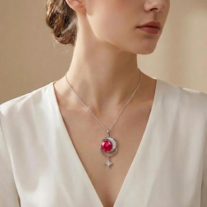 May include: Silver necklace featuring a pendant with a large, round, ruby-red gemstone. The pendant is set within a crescent moon design, embellished with small, clear crystals. A star-shaped charm dangles below the moon, also adorned with crystals.