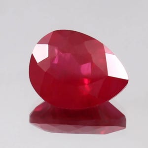 May include: A pear-shaped, faceted gemstone with a deep, vibrant red color. The stone features multiple triangular and trapezoidal facets that reflect light, creating a brilliant, translucent appearance with a clear reflection visible below.