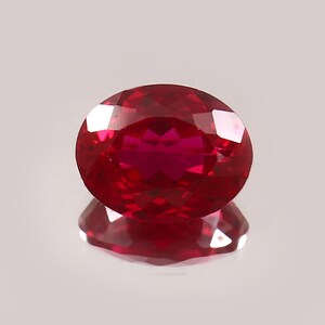Può includere: Una gemma di rubino sfaccettata a forma ovale. La gemma è di un colore rosso intenso e saturo con riflessi interni. Il rubino è posto su uno sfondo chiaro, con il suo riflesso visibile sotto. La pietra preziosa è un punto focale, che mostra la sua chiarezza e brillantezza.