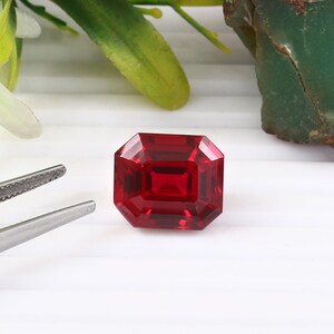 May include: A faceted, rectangular-cut, deep red gemstone. The gem has a square shape with beveled edges, reflecting light. The stone is set against a white background, with a pair of tweezers and greenery in the background.