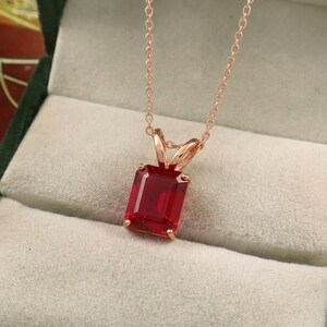 May include: A rose gold necklace featuring a rectangular, deep red pendant. The pendant is a large, faceted gemstone, suspended from a delicate chain. The necklace is displayed in a soft, neutral-toned jewelry box, highlighting the pendant's color.
