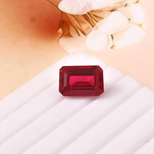 May include: A rectangular, faceted, deep red gemstone. The gem is set against a white, ridged surface, with a soft, peach-coloured background. The gem's facets reflect light, creating a brilliant appearance.