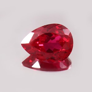 Könnte beinhalten: Ein facettierter, birnenförmiger Rubin-Edelstein. Der Edelstein ist ein tiefes, leuchtendes Rot mit einer polierten Oberfläche, die das Licht reflektiert. Der Rubin liegt auf einer reflektierenden Oberfläche und erzeugt einen Spiegeleffekt. Symbol für Leidenschaft und Liebe.