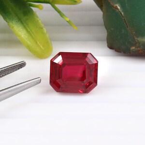 May include: A faceted, rectangular-cut ruby gemstone with a deep red hue. The gem is displayed on a white surface, with a pair of tweezers and green foliage in the background. The ruby's facets reflect light, showcasing its clarity and brilliance.