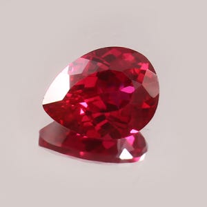 Könnte beinhalten: Ein facettierter, birnenförmiger Rubin-Edelstein. Der Edelstein hat eine tiefe, gesättigte rote Farbe mit mehreren Facetten, die das Licht reflektieren. Der Rubin liegt auf einer reflektierenden Oberfläche.