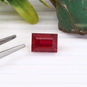May include: A rectangular, faceted, deep red gemstone. The gem is set against a white background, with a pair of silver tweezers to the left. Green foliage and a piece of green stone are in the background.