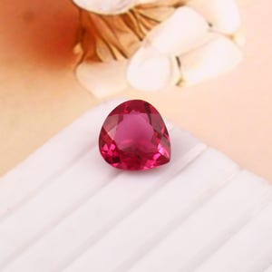 May include: A faceted, heart-shaped, ruby-red gemstone. The gem is set on a white, ridged surface, with a soft, blurred background of cream and tan hues. The gemstone's facets reflect light, creating a sparkling effect.