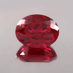 May include: A faceted, round-cut ruby gemstone. The gem is a deep, saturated red color, with multiple facets catching the light. The ruby is displayed on a reflective surface, creating a mirrored effect. The gemstone is a symbol of love and passion.