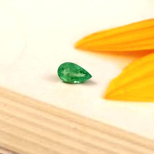 Pode incluir: Uma gema de esmeralda verde vibrante em forma de pêra. A gema está sobre uma superfície de madeira clara, com uma pétala amarela desfocada no fundo. As facetas da esmeralda capturam a luz, mostrando sua clareza e cor.