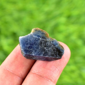 May include: A rough-cut, natural blue sapphire gemstone. The sapphire has a dark blue color with some areas appearing almost black. The edges have a brownish hue. The gemstone is held in a person's hand against a blurred green background.