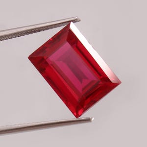 May include: A vibrant, rectangular-cut ruby gemstone held by silver tweezers. The ruby displays a deep red hue with facets that catch the light, showcasing its clarity and brilliance. The gemstone is a focal point against a neutral background.