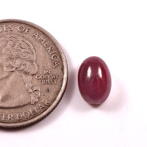 May include: A polished, oval-shaped, deep red gemstone next to a U.S. quarter. The gemstone has a smooth, glossy surface. The quarter shows the profile of George Washington and the words "In God We Trust".
