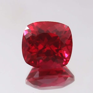 Puede incluir: Un rubí tallado en cojín, que muestra un color rojo intenso y una faceta intrincada. Las facetas de la gema capturan y reflejan la luz, creando un brillo brillante. El rubí se presenta sobre una superficie reflectante.