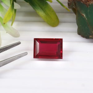 以下が含まれることがあります： 深紅色の長方形のファセットルビーの宝石。白い背景に配置され、ピンセットと緑の植物が見えます。ルビーは鮮やかで人目を引くジュエリーです。
