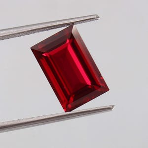 May include: A vibrant, deep red gemstone cut into a rectangular emerald shape. The stone features sharp, clean facets that reflect light, creating a brilliant, saturated appearance. It is held securely by a pair of silver-toned metal tweezers.