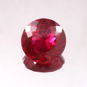 May include: A faceted, round, deep red gemstone with a brilliant cut. The gem reflects light, creating a sparkling effect. The gem is set against a white background, with its reflection visible below.
