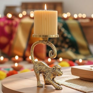 May include: A gold-colored monkey candle holder with a lit, beige pillar candle. The monkey is in a crawling position, supporting the candle holder with its curved tail. The candle holder has a decorative, swirled design.