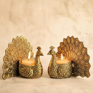 May include: Two brass peacock candle holders with lit candles. The candle holders are detailed with feathers and have a gold finish. The candle holders are designed to hold small votive candles.