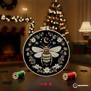 Puede incluir: Un bastidor de bordado negro con un diseño detallado de abeja, una luna creciente y detalles florales. La abeja tiene intrincados detalles de alas y cuerpo. El bastidor está rodeado de patrones florales y de estrellas blancos y dorados. Dos carretes de hilo y botones rojos están sobre la mesa.