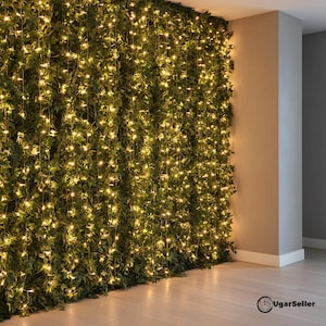Op de afbeelding: Een muur bedekt met weelderig groen gebladerte, verlicht door strengen warme witte feeërieke lichtjes. De lichten vallen naar beneden, waardoor een zachte, uitnodigende gloed ontstaat. De muur staat tegen een neutraal getinte muur en een lichtgekleurde houten vloer.