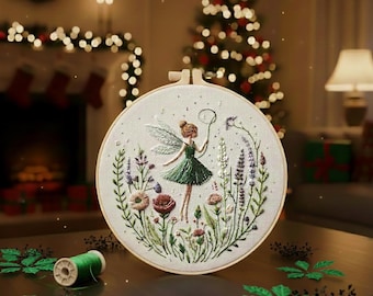Könnte beinhalten: Gestickte Feen-Hoop-Art mit einer Fee in einem grünen Kleid mit floralen und pflanzlichen Details. Die Fee hält eine Schleife. Die Stickerei ist in einem Holzreifen angebracht. Der Hintergrund umfasst einen Weihnachtsbaum und einen Kamin.