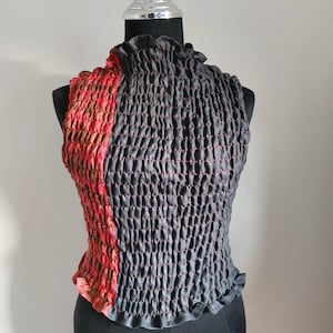 Op de afbeelding: Mouloos topje met een hoge halslijn, weergegeven op een mannequin. De top heeft een getextureerd, gesmokt ontwerp in donkergrijs met rode stiksels. Eén kant is een levendig rood-bruin patroon.