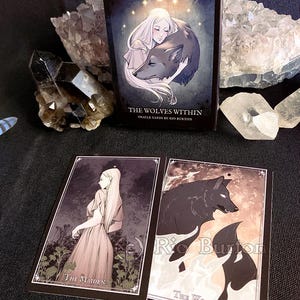 May include: A box of oracle cards titled "The Wolves Within" by Rio Burton. The box features a woman with long white hair holding a wolf. The cards are illustrated with a woman and a wolf.