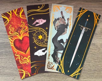 May include: Four fantasy-themed bookmarks. One features a red heart with flames, another has hands and a star, a third depicts a wolf, and the last shows a sword. Each bookmark has gold accents and a unique background color.