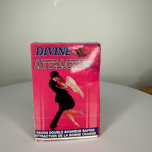 Puede incluir: Una caja de jabón rosa con las palabras "DIVINE ATTRACTION" en una fuente estilizada. La caja presenta una ilustración de una pareja abrazándose, con una persona con alas de ángel. También está presente el texto "SAVON DOUBLE BONHEUR RAPIDE ATTRACTION DE LA BONNE CHANGE".