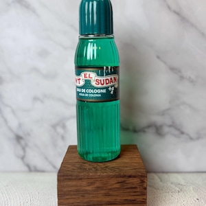 Puede incluir: Una botella de vidrio verde vintage de "El Sudan" eau de cologne. La botella tiene un diseño acanalado y una tapa verde oscuro. La etiqueta es negra, roja y blanca. La botella se exhibe en un pequeño bloque de madera oscura.