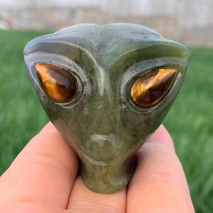May include: A green alien head sculpture with large, oval, tiger's eye gemstone eyes. The sculpture is held in a hand, with a blurred green background.