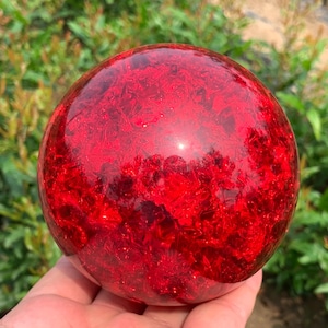 Puede incluir: Un objeto esférico liso hecho de material rojo translúcido con un interior cristalino texturizado. La superficie está pulida y es reflectante, mostrando refracciones de luz internas y tonos carmesí profundos en toda la forma.